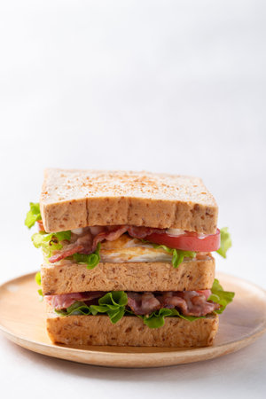 Whole wheat bread sandwich with bacon, lettuce, tomato and fried egg on wooden plate with white backgroundの写真素材