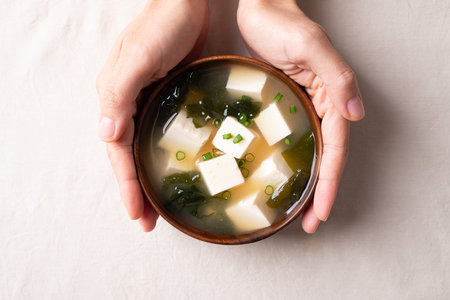 Top view of hand holding healthy Japanese miso soup with soft tofu and seaweed in a wooden bowl on white fabric backgroundの写真素材