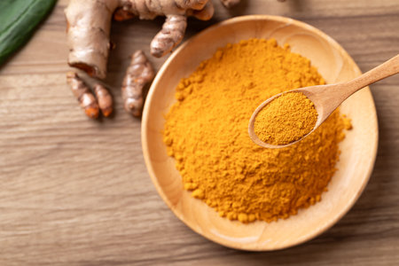 Top view of turmeric powder in a wooden bowl with a wooden spoon scooping it, and fresh turmeric...
