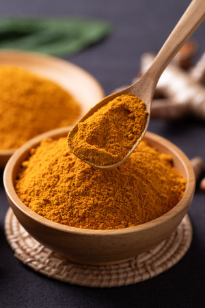 Turmeric powder in a wooden bowl with a wooden spoon scooping it, and fresh turmeric roots on a...