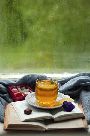 A cup of tea with lemon, books and candies on the background of a window on a rainy day.の写真素材