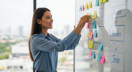 Young businesswoman writing on sticky notes on glass board in office. She is looking at sticky notes on whiteboard and smiling.の素材