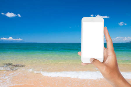 Hand holding smartphone with beautiful scene, tropical sea and beach with blue sky background