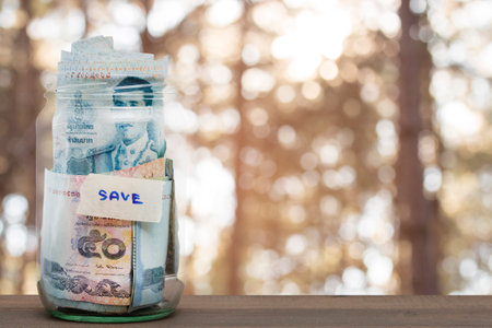 money in the glass jar on blurred leaf background. business concept of saving and investment.の写真素材