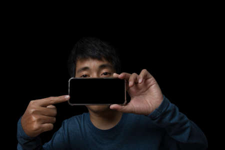 Young man holding smartphone pointing finger at blank screen on black background.の写真素材