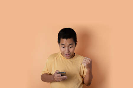Portrait of young man standing using mobile phone make winner gesture on orange background.の写真素材