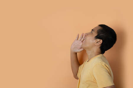 Young man shouting and screaming loud to side with hand on mouth isolated on orange studio background.の写真素材