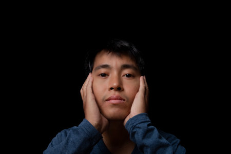 Portrait of a young man wearing blue t-shirt touching cheek by hand looking at camera studio shot on black background. copy spaceの写真素材