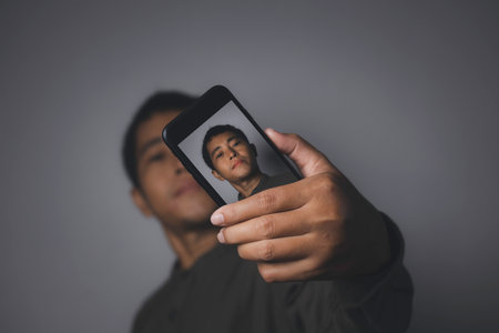 Young man using smart phone shooting selfie back camera. Portrait of man having video-call with smart phone. Human emotion face expression concept. Studio shot isolated on gray background enjoying.の写真素材