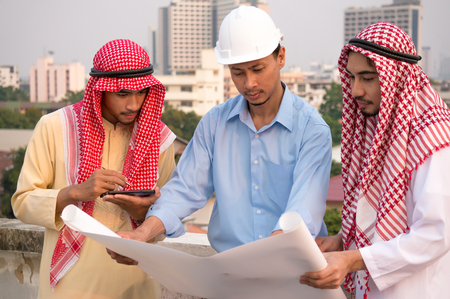 Two Arab businessman and engineer wearing safety helmet holding paper discussing about project, construction and investmentの写真素材