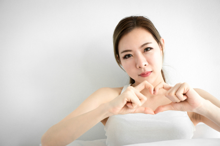 Asian beauty woman show hand heart on white bed in white bedroom in morningの写真素材