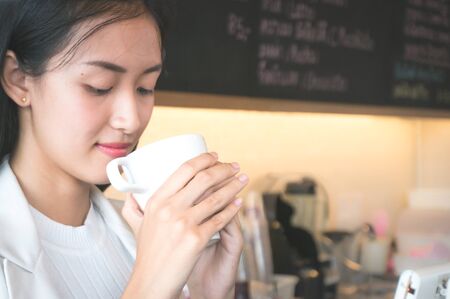 Asian woman hold and drink coffee white cup in hand in coffee shop in morningの写真素材