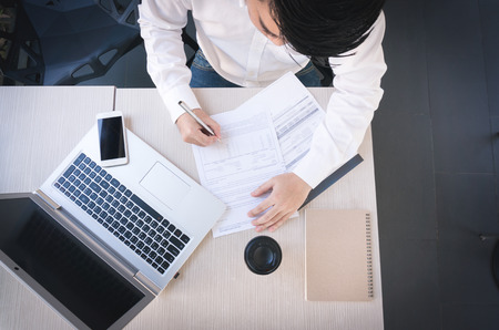 A business man in white shirt holding pen in hand to sign contract on table with laptop, coffee cup, notebook and smart phone in coffee shop by top viewの写真素材
