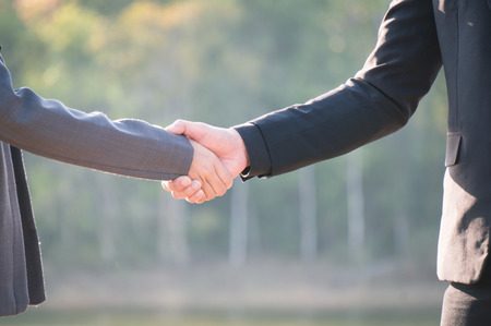 Two engineers wearing formal suit and safety helmet shake hands on riverside among sunlight in national parkの写真素材