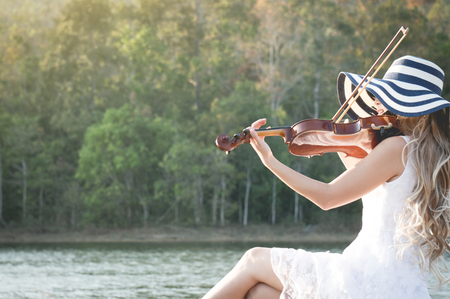 A woman / violinist wearing white dress and hat playing violin on riverside among sunlight feeling freedomの写真素材