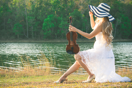 A woman / violinist wearing white dress and hat playing violin on riverside among sunlight feeling freedomの写真素材
