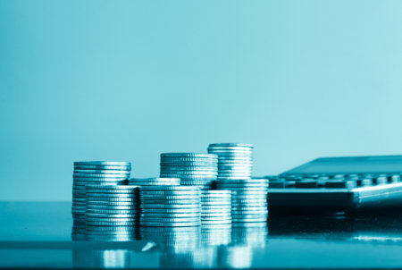 Coins stacked on each other with calculator and pen on blue backgroundの写真素材
