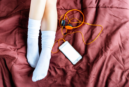 Charming beautiful woman is lying down on a bed with her smartphone or mobile phone, headphone. Attractive woman wears long white socks in the winter time at bedroom with white, isolated screen phoneの写真素材