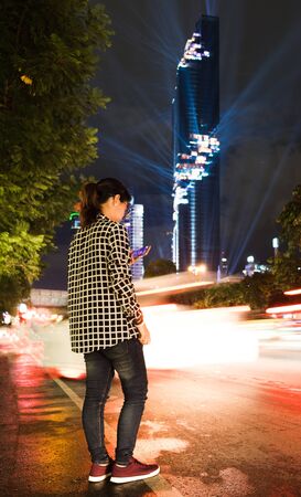 young woman standing playing smart phone in city night time have mahanakhon tower on backgroundの写真素材