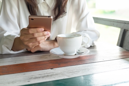 front view. young woman playing mobile phone while take a break in morning time. front of her having coffee cup putting on wooden table. this image for business, technology, beverage and portrait conceptの写真素材