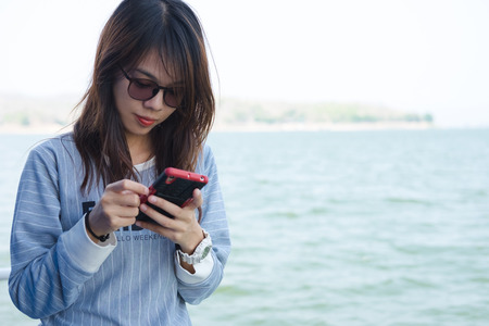 lonely young woman playing mobile phone while standing and have blue sea are background. this image for nature,portrait and travelの写真素材
