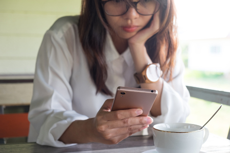 front view. young woman playing mobile phone. she make serious face while take a break in morning time. front of her having coffee cup putting on wooden table. this image for business, technology, beverage and portrait conceptの写真素材