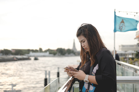 young asia woman stand alone on riverside and she looking smart phone on her hand  with scenery view are background. this image for person,portrait,nature,city,travel,building conceptの写真素材