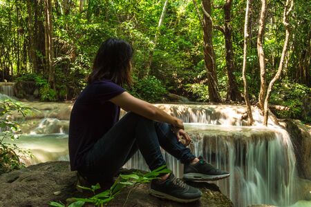 young asia woman sitting alone on the rock among the forest with beautiful waterfall are background. image for nature, forest, landscape, portrait, wild, person conceptの写真素材
