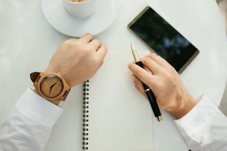 business background hand of businessman wearing wooden watch with pen, notebook, coffee, mobile phone on table. image for technology, education, fashion, accessory, equipment, document, idea conceptの写真素材