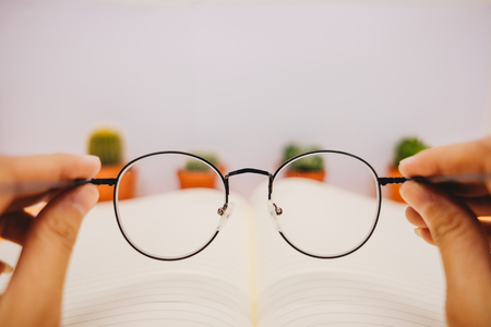 front view hand of businesswoman holding glasses with blank paper and cactus place on table background. image for copy space, desktop, workplace, workspace, education, plant, isolated, defocus conceptの写真素材