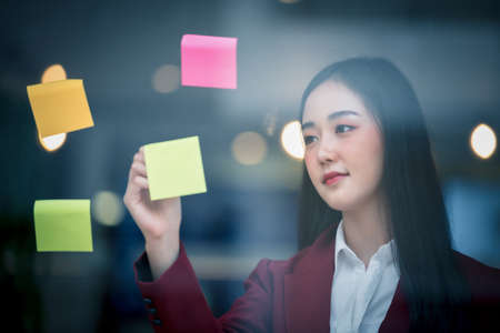 Portrait of Young asian businesswoman writing sticky paper pad on glass in modern office. Business...