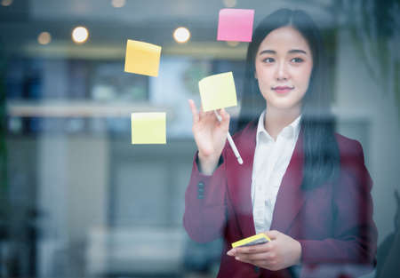 Portrait of Young asian businesswoman writing sticky paper pad on glass in modern office. Business...