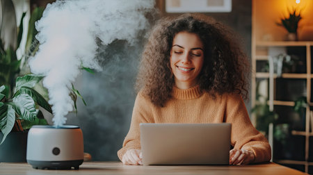 Essential oil diffuser emitting steam, young woman smiling while working on laptop, peaceful and productive home office setup.の素材