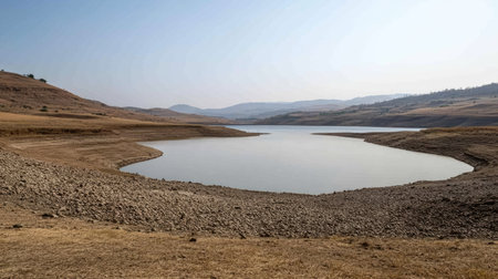 Still water of Bangwad Dam during dry season, revealing rocks and shoreline textures under clear skyの素材