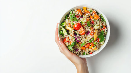 A hand holding a bowl of colorful quinoa salad with fresh organic vegetables, captured from above on a clean white background, perfect for a healthy vegan meal.の素材