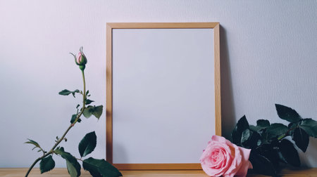Blank frame poster leaning against the wall on a table, with a delicate pink rose beside it. A chic and simple scene for design mockups or decor inspiration.の素材