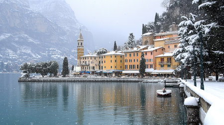 Picturesque Riva del Garda town under a light snowfall, with the tranquil waters of Garda Lake and surrounding snow-dusted mountains creating a winter wonderland.の素材