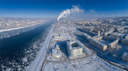 This stunning aerial shot captures a winter cityscape featuring a frozen river, snow-covered buildings, and industrial smoke against a clear blue sky.の素材