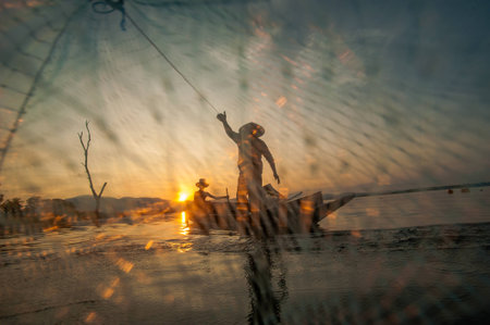 Asian fisherman on wooden boat casting a net for catching freshwater fish in nature river in the early morning before sunrise.Fishermen fishing in the morning light.の写真素材