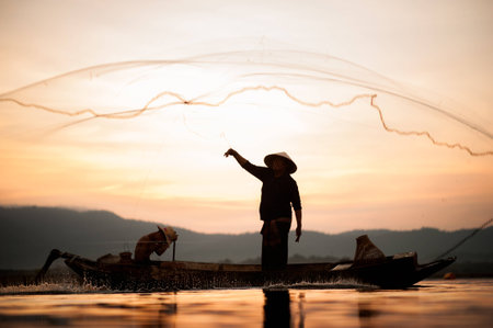 Asian fisherman on wooden boat casting a net for catching freshwater fish in nature river in the early morning before sunrise.Fishermen fishing in the morning light.の写真素材