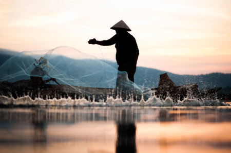 Asian fisherman on wooden boat casting a net for catching freshwater fish in nature river in the early morning before sunrise.Fishermen fishing in the morning light.の写真素材