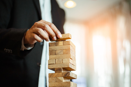 business man hand  pulling out or placing wood block on the tower in modern office. plan and strategy in business. blur for background.の写真素材