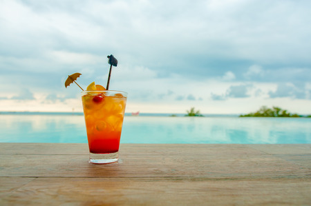 Cocktail at the edge of the swimming pool with sky. Red cocktail with an orange slice on the background of the swimming pool and sky.の写真素材