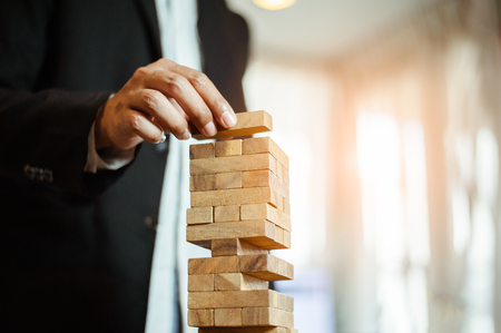 business man hand  pulling out or placing wood block on the tower in modern office. plan and strategy in business. blur for background.の写真素材
