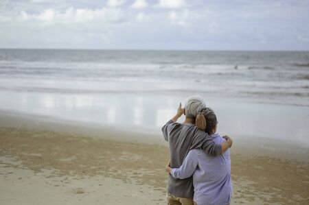 A senior couple hugged each other at the beach in the morning. The old man pointed the finger to the old woman to look at the bright blue sky, life insurance plan at the retirement concept.の写真素材