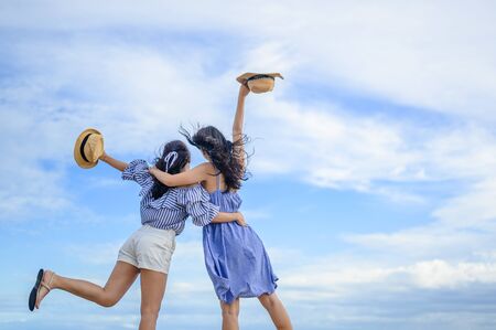 Two happy woman holding straw hat with hand raised to sky.の写真素材