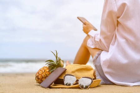 Girl texting in a smartphone on the beach With a hat, sunglasses, passport and pineapple.Technology and relaxation comcept.の写真素材