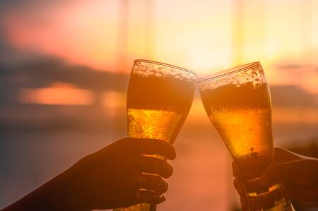 Couple hands holding beer gass and clanging, at the bar in summer sunsetの写真素材