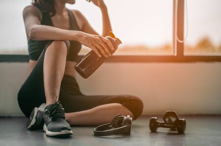 Fitness woman Relaxing after exercise with a whey protein and dumbbell placed beside the gym.Relaxing after training.beautiful young woman looking away while sitting  at gym.の写真素材