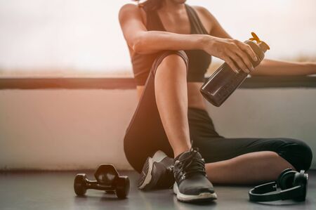 Fitness woman Relaxing after exercise with a whey protein and dumbbell placed beside the gym.Relaxing after training.beautiful young woman looking away while sitting  at gym.の写真素材